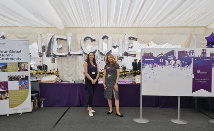 Two staff members standing in front of an alumni stand at graduation with balloon welcome sign