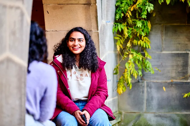 Two students sitting in the courtyard of St Marys on a sunny day having a friendly conversation while sitting and leaning against limestone buildings