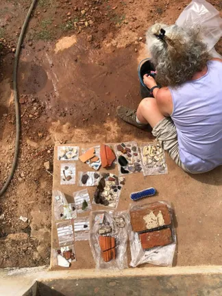 Top down photo of a person with curly silver and brown hair, lavender tank top and beige shorts, sitting on a step and cleaning an archaeological find over a bucket. On their left is laid out an array of archaeological finds on top of small plastic bags. The visible ground is reddish brown with dry soil and sparse grass.