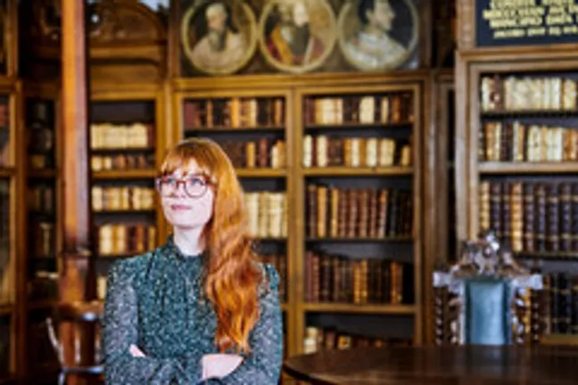 Female student/intern with long red hair and glasses standing in front of books inside a glass and wood display cupboard