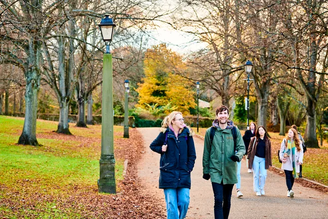 Male and female students walking along path with autumn trees in the background and leaves on the ground