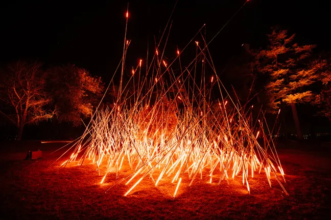 An artwork made up of long bamboo poles lit up red to give the impression of fire and energy, set in an open space at night-time.