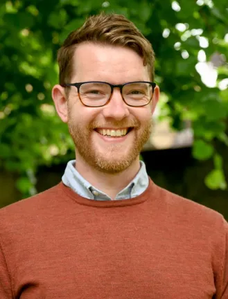 Headshot of David Lowther, he is light skinned with brown hair wearing glasses and a brown jumper.