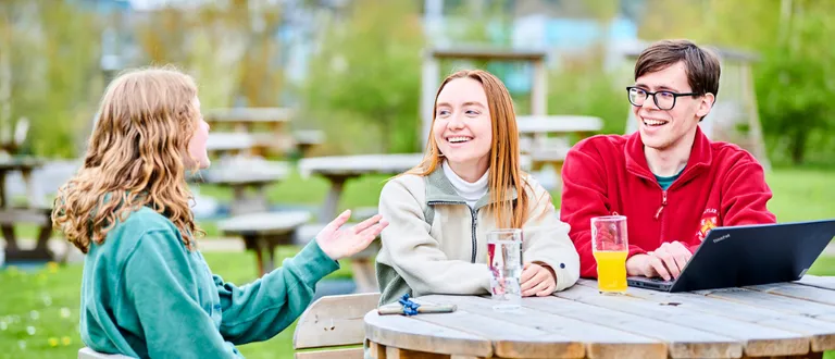 Three students sit outside at a wooden picnic table on the lawn in front of Josephine Butler College on a sunny day