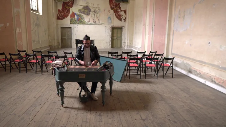 A young man tuning a piano in a large room with another piano and chairs laid out behind, as though for a concert; stillshot from Hopa Lide film, directed by Petr Nuska