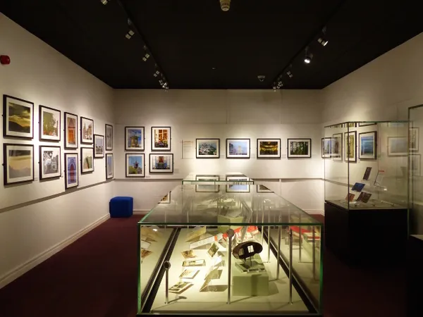 A rectangular gallery with white walls, black ceiling and red carpet.  Around the walls are colourful photographs of landscapes in white mounts and black frames. Down the centre of the gallery are two display cases with many objects inside, another taller display case is visible on the right.