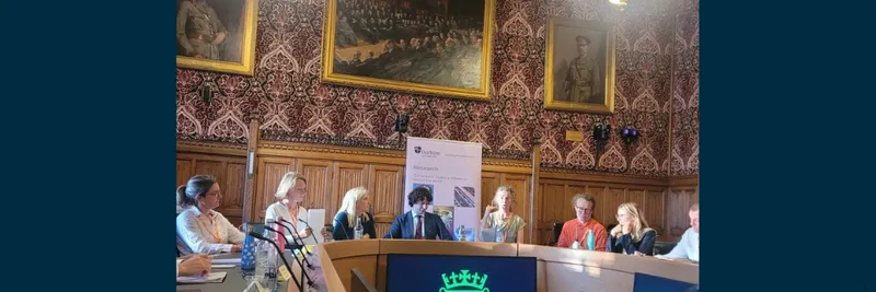 Panel of speakers sat at horseshoe shaped table in large room within the Houses of Parliament