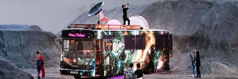 A single decker bus spray painted with images of space and featuring a satellite dish on its roof is parked in a dusty, moon-like landscape.