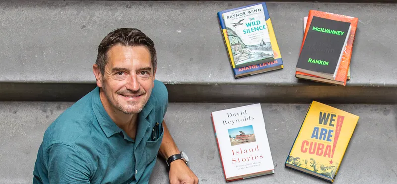 A smiling man sitting on some stairs surrounded by books