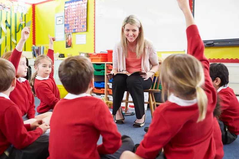 Female primary school teacher sat on chair with children sat on the floor round her. Some have their hands raised.