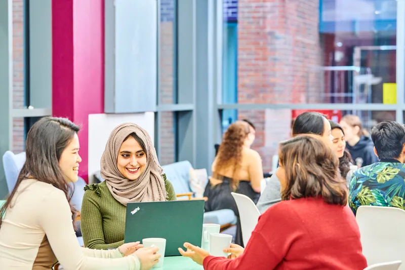 Group of postgraduate students smiling around a laptop with mugs