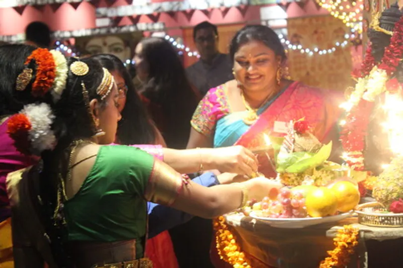 A lady dressed in traditional Indian clothing presides over Lakshmi Puja while another person gives offerings to the Lakshmi Puja shrine