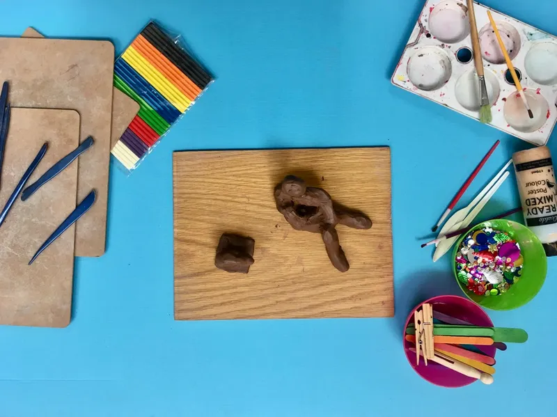 Aerial view of blue table with wooden board in centre. On the board are two pieces of brown clay that have been sculpted in to Henrey Moore style sculptures.