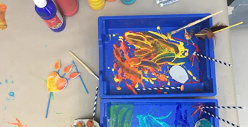 Downwards view of a table covered with bottles of paint, two blue trays filled with different colours of paint swirled together with paint brushes and straws lying in them and a completed artwork made of straws and card in the shape of a heart.