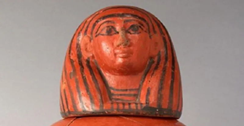 Close up view of the lid and upper section of a canopic jar featuring the human headed god Imsety, made of orange clay with details picked out in a black glaze.