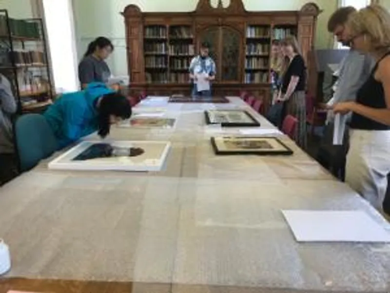 A group of students are standing around a table in the centre of the room, which is covered in bubble wrap. On top of this are framed images of artworks, which the group is looking at. One of the group has leaned over the table for a closer look.