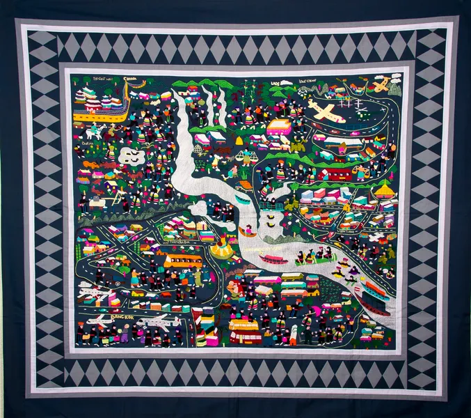 Photograph of a square fabric banner known as a story cloth. It is covered with scenes on a blue background showing the history of the Hmong people of South East Asia, who are depicted in traditional clothes of black with red belts.
