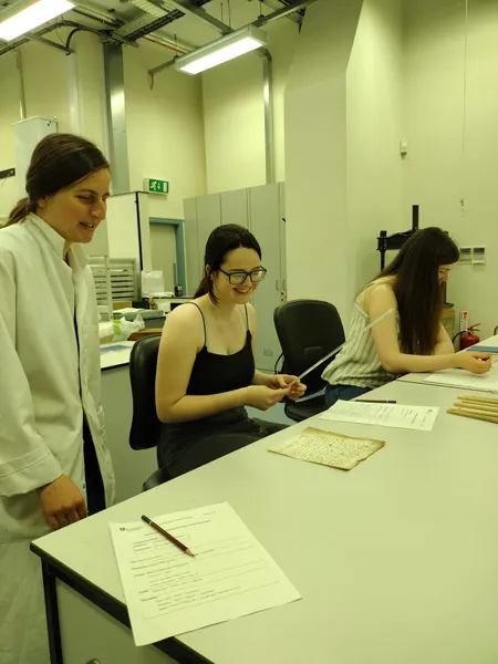Two Work Experience participants sit at a table in a Conservation studio with historic documents and modern forms on the table. A member of the Conservation team stands next to them smiling.