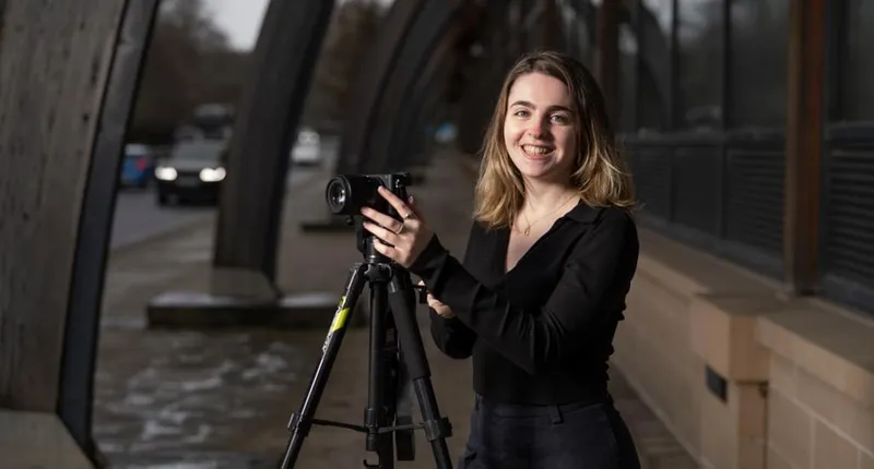 A smiling student stands outside Durham University's Palatine Centre, with her hands on a video camera on a tripod.