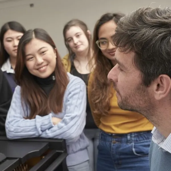 A group of students listening to a lecturer play the piano