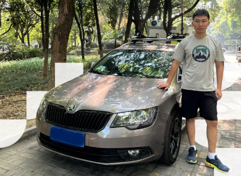 A young man in shorts, t-shirt and trainers standing to the right of a silver car with his hand on the bonnet