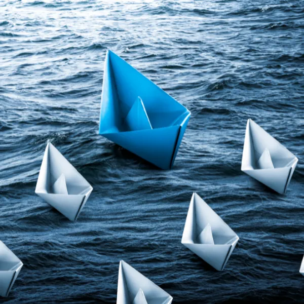 A blue paper sailing boat on water followed by smaller white paper boats
