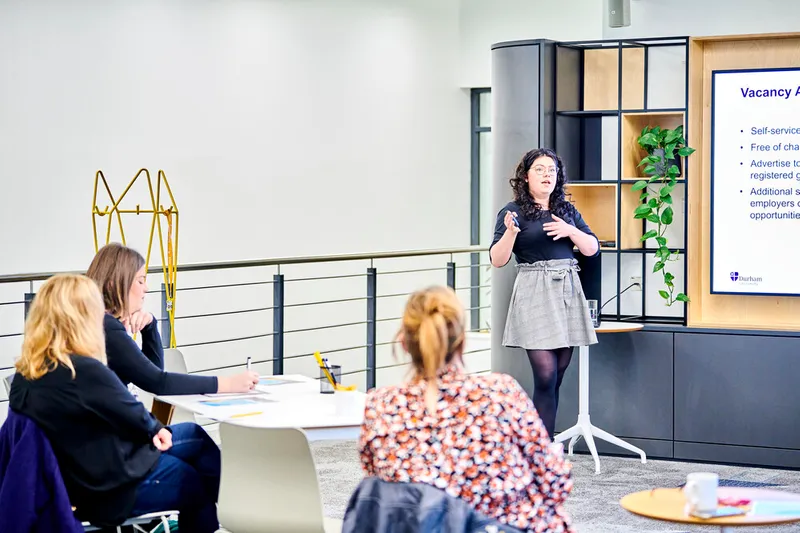 A staff member talks to a group of people. She is standing next to a digital screen and holds a remote control in her hand. Attendees sit at a table listening and making notes