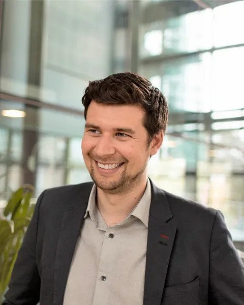 A brown haired man with a shirt and jacket smiling off camera