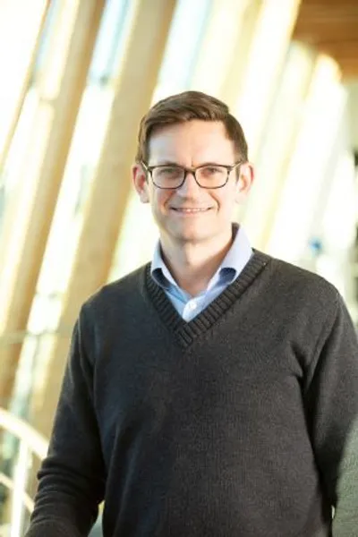 A man wearing a dark v-neck jumper and glasses smiling at the camera