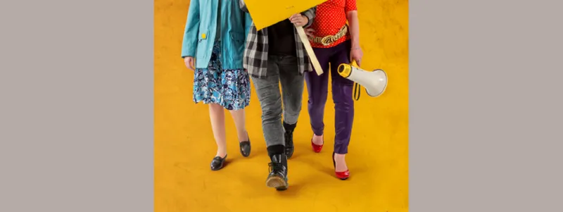 Three women pictured from the waist down walking towards the camera carrying a placard and a megaphone