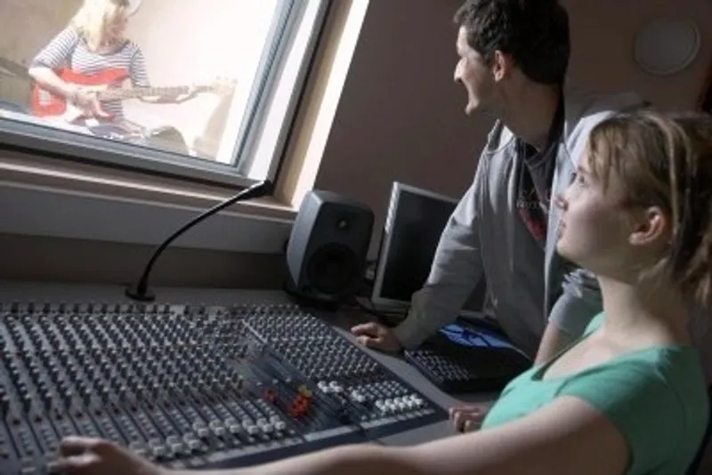 Students in a recording studio booth sitting at a large mixing deck