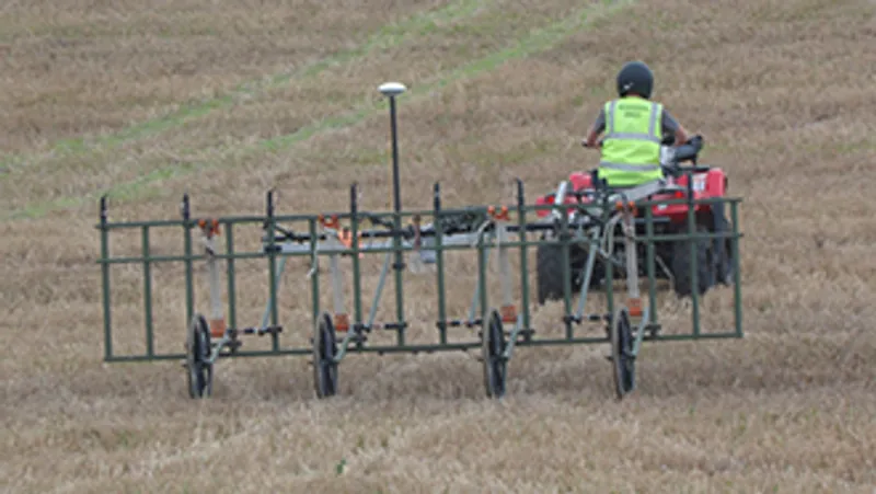 A person riding a quad bike across a field, towing an array of geophysical sensors on a mobile cart