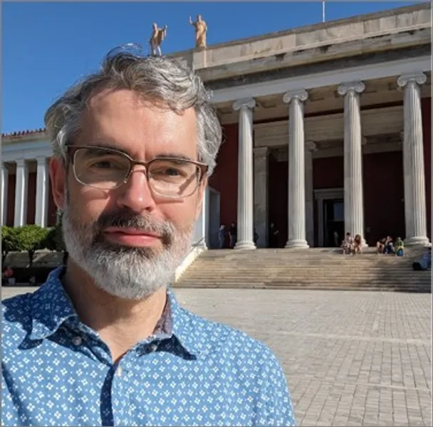 Dr Rob Witcher smiling and standing in front of some Classical architecture under a blue sky on a sunny day.