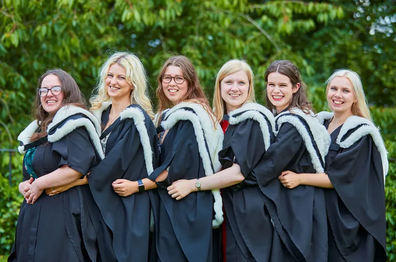 Six women stand in a row, hugging each other around the waist and smiling; they are wearing graduation outfits of black robes and black hoods with white fur trim