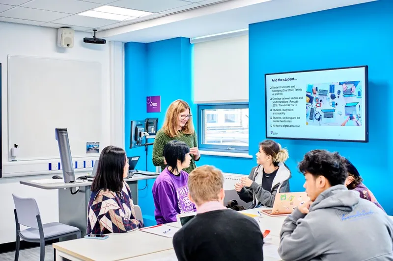 Brightly lit, contemporary seminar room with multiple screens on the walls and at the front of the room. Students sit together at trestle tables, and a female academic in a green dress is talking to them and using the screens to share her research