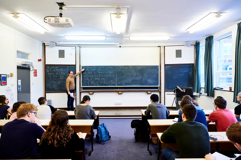 Students sitting in class while the tutor points to the blackboard.