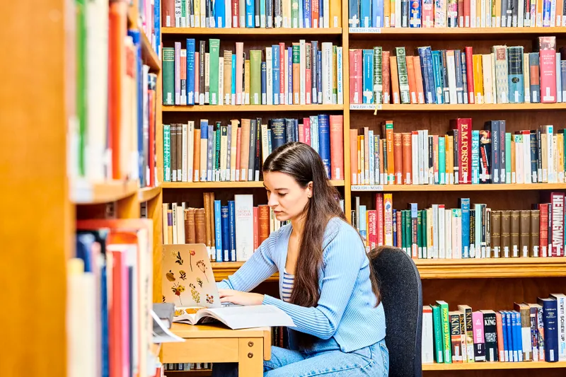 Student looking at book in front of bookshelf