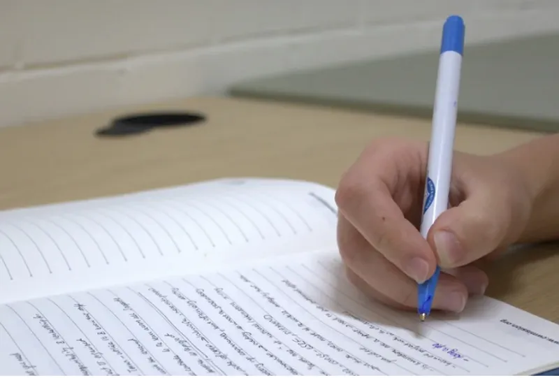 A hand holds a pen that is writing a report on lined paper