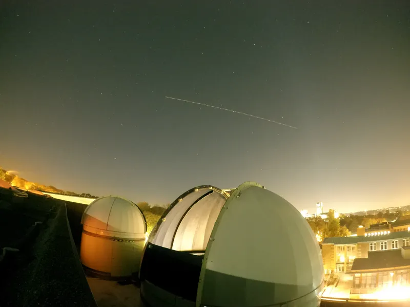 Night time shot of telescope domes on the roof of the building and a shooting star above. The cathedral can be seen lit up in the distance.