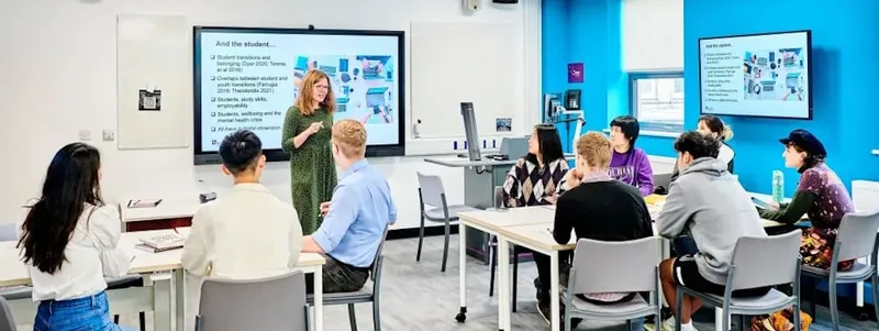 Brightly lit, contemporary seminar room with multiple screens on the walls and at the front of the room. Students sit together at trestle tables, and a female academic in a green dress is talking to them and using the screens to share her research