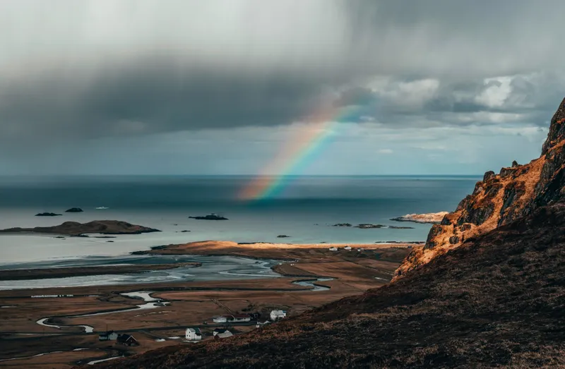 A panorama over a cold looking, brown landscape with small white buildings and grey/blue waterways finishing up in the sea. There is a rainbow ending in the sea.