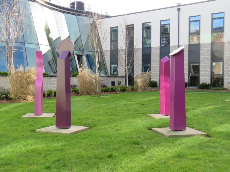 Four purple pillars of slightly different shapes and sizes stand on stone bases arranged in a group on a lawn with a building in the background