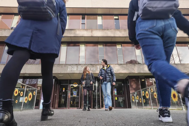 Two people walking out of Bill Bryson Library framed by two other people walking towards the building.