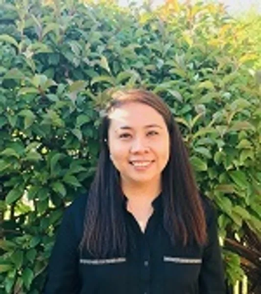 Headshot of Mel Earnshaw, she is brown skinned and wearing a black shirt and is standing on front of a leafy hedge.