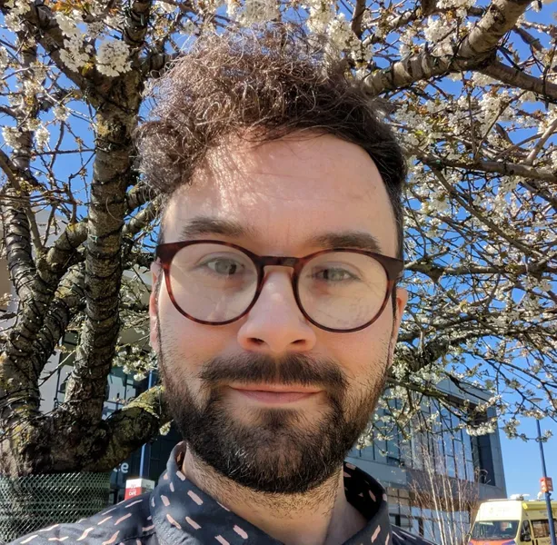 Andrew is light skinned with brown, curly hair and a brown beard. Andrew is wearing glasses and a navy and pink shirt. In the background are blue skies and a blossom tree