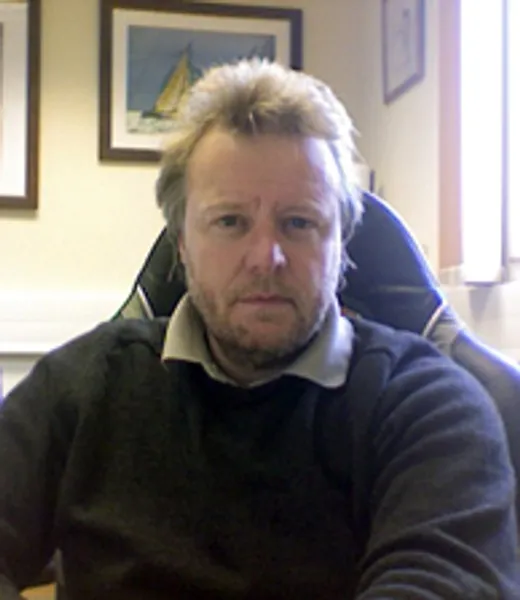Professor Andy Monkman, BSc., PhD., C.Phys, FInstP sitting in a desk chair in front of framed photographs