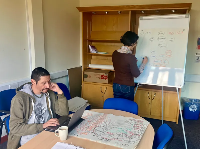 Person sat at table working on computer with person standing writing on whiteboard