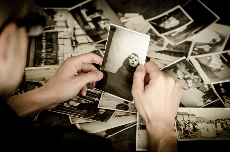 A close-up image of someone's hands holding an old photograph showing a young girl, whilst behind can be seen many old black and white photos, scattered across a table surface