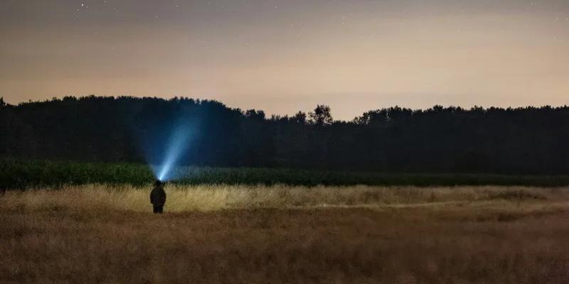 Person standing in a field with trees on the horizon. The person is looking up at the night sky and there is is faint beam of blue light emitting from their head.
