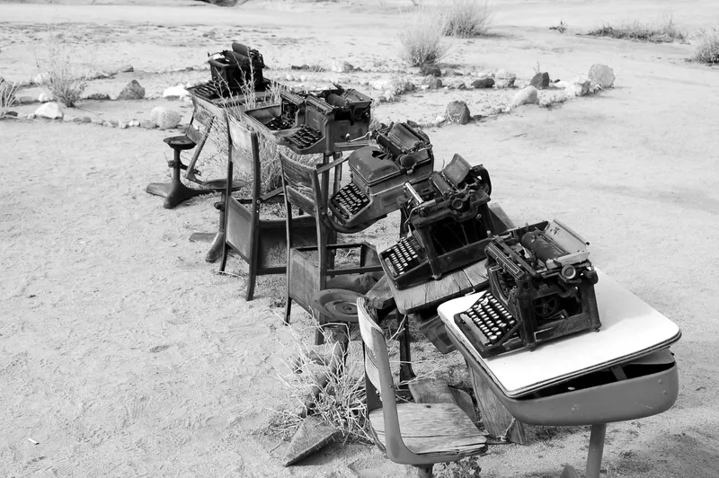 Black and white photo of line of desks on beach with typewriters and chairs